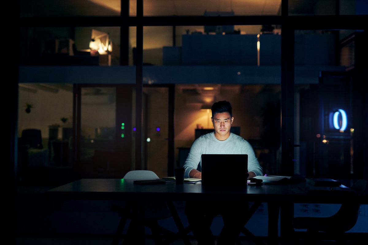 Mann arbeitet abends allein im dunklen Büro am beleuchteten Laptop